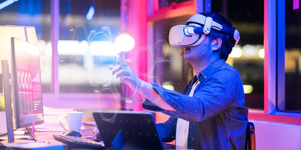 A person using a virtual reality headset while interacting with holographic projections at a desk equipped with a laptop, monitor, and cup, all set in a vibrant, neon-lit environment reminiscent of the metaverse.