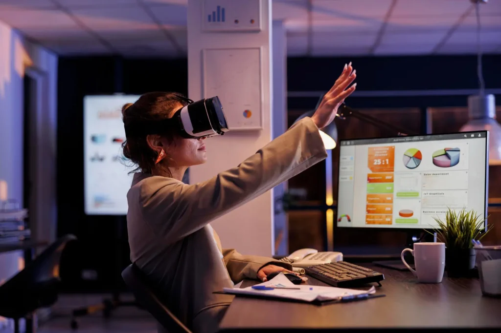 A person wearing a VR headset uses their hand to interact with the virtual reality environment, while sitting at a desk with a computer screen displaying charts and graphs.